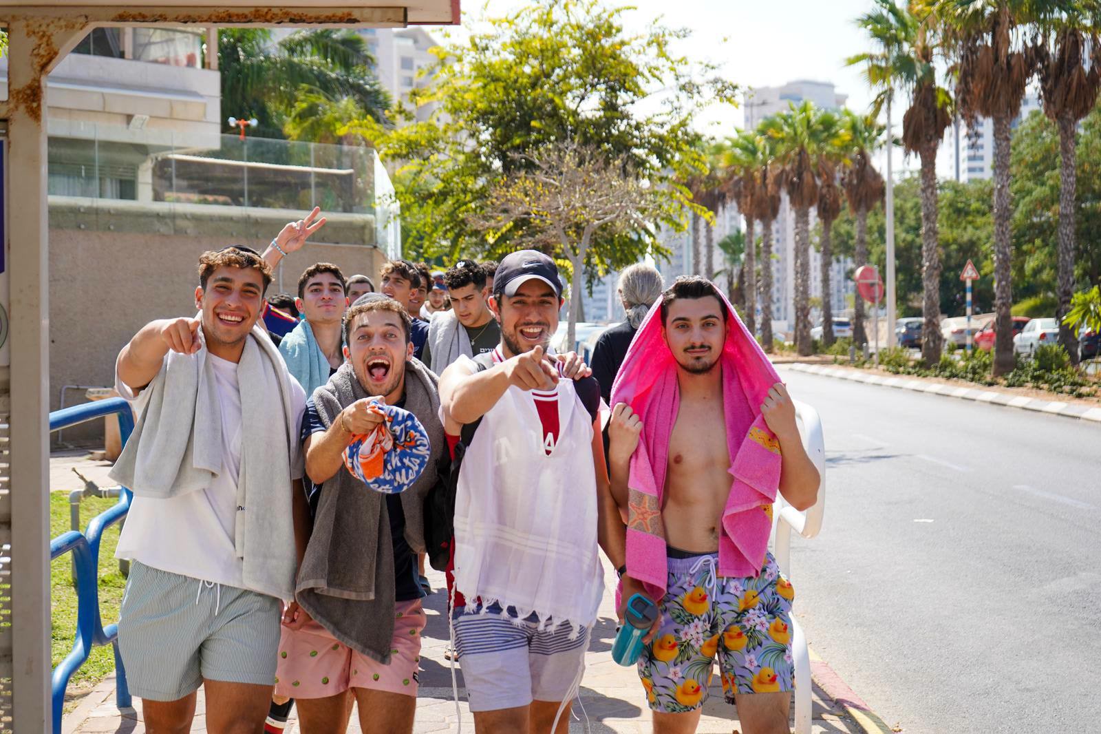 Young men with beach towels over their shoulders, laughing and pointing at the camera on a palm-lined street.